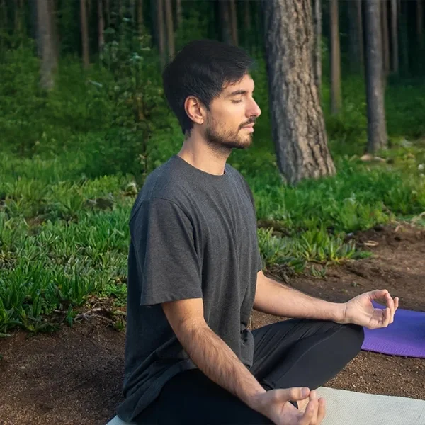 Hombre meditando con los ojos cerrados sobre un tapete de yoga en un bosque rodeado de árboles y vegetación.