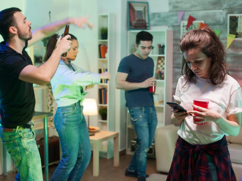 Cheerful young woman sitting in the centre of living room using her smartphone. Young people dancing.